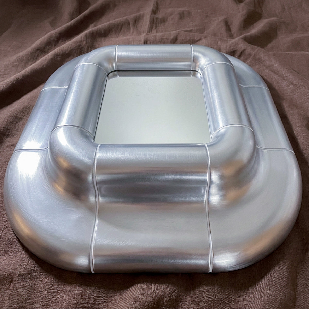 Close view of the Lolo Steel mirror on brown fabric, showing the cushioned frame and brushed metallic lacquer