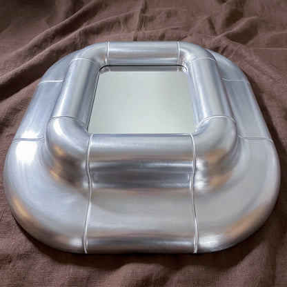 Close view of the Lolo Steel mirror on brown fabric, showing the cushioned frame and brushed metallic lacquer